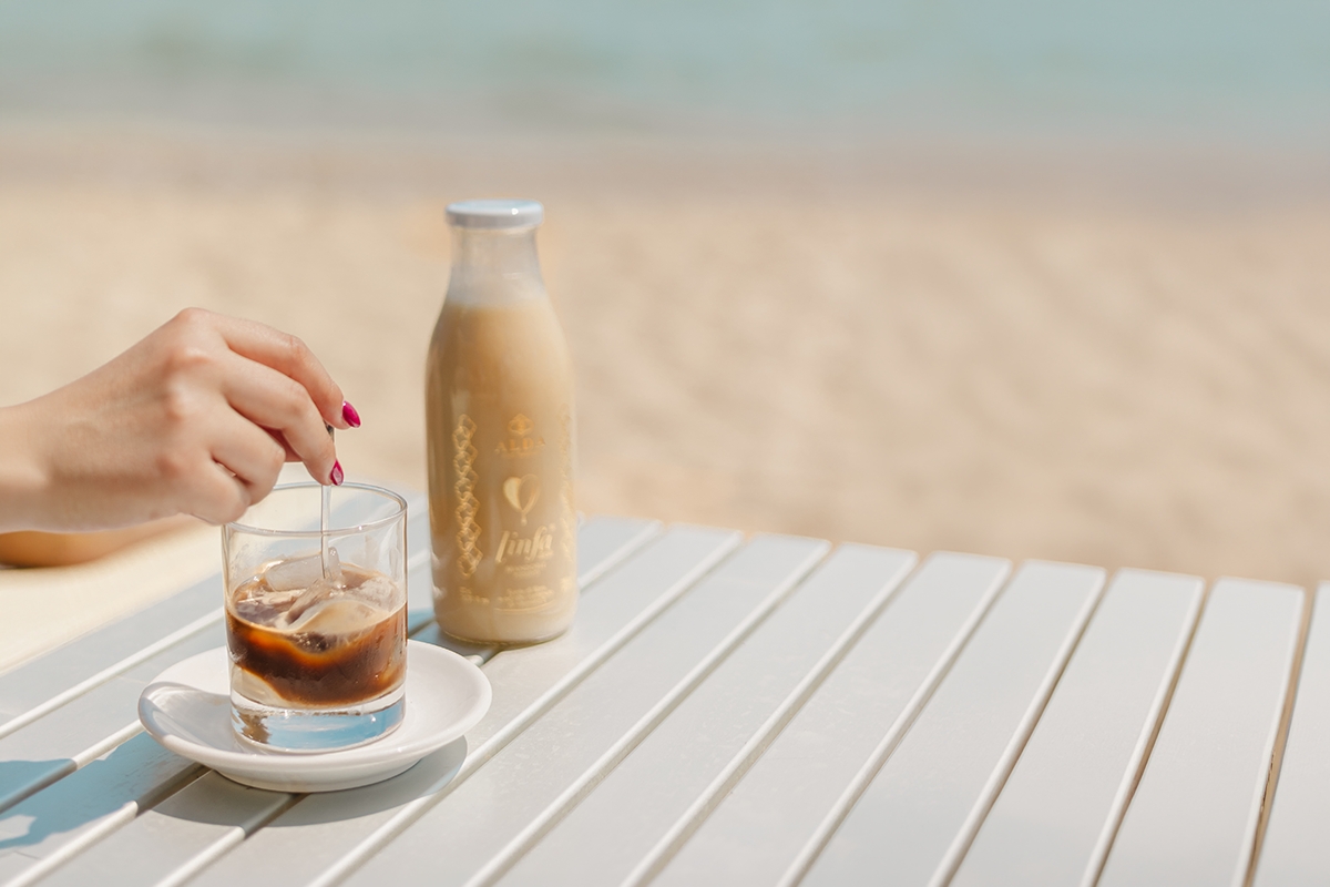 Foto di un bicchiere di caffè in ghiaccio con linfa di mandorle e bottiglia di linfa con uno sfondo di sabbia e mare